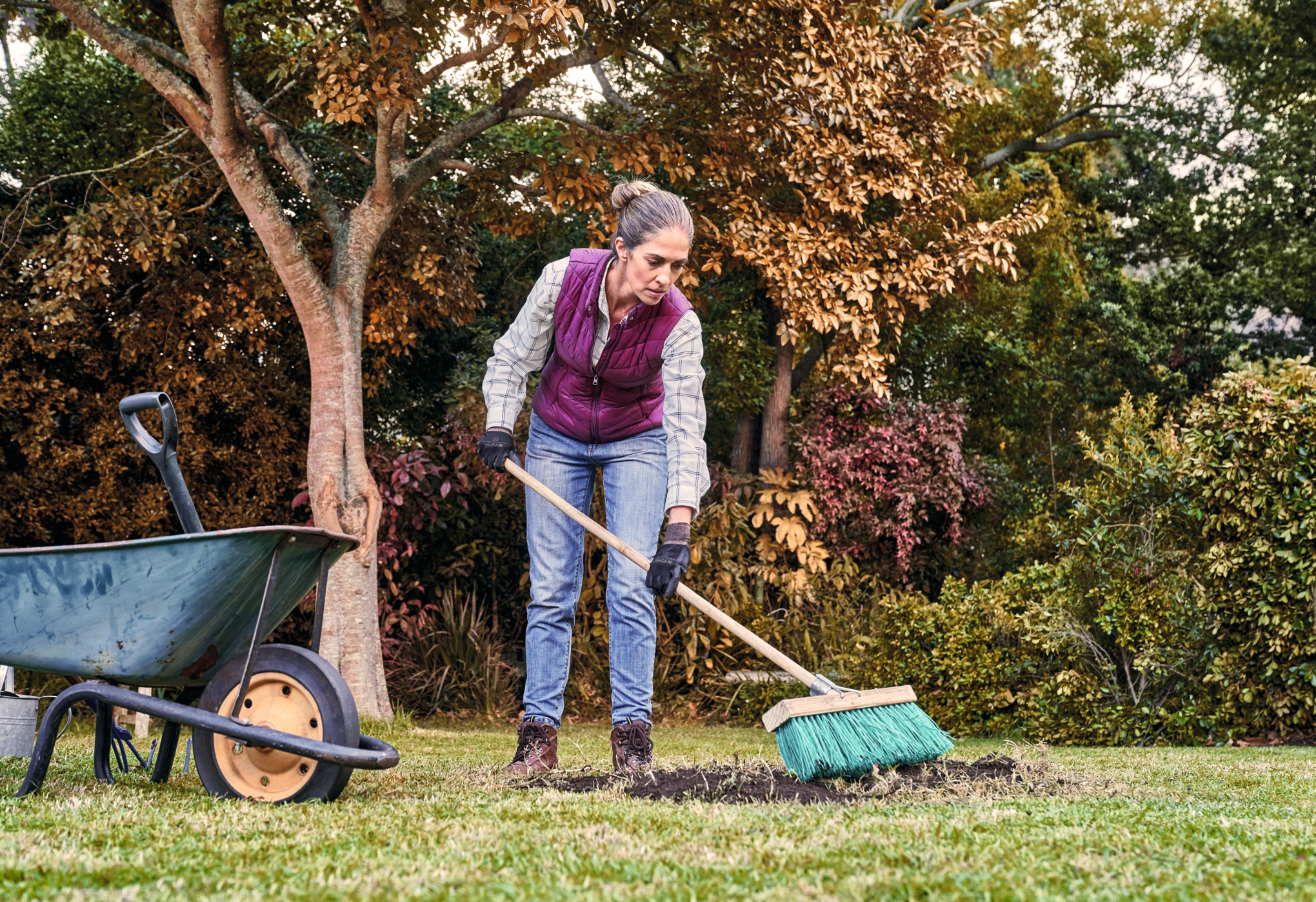 A woman sweeps leaves in a garden beside a metal wheelbarrow.