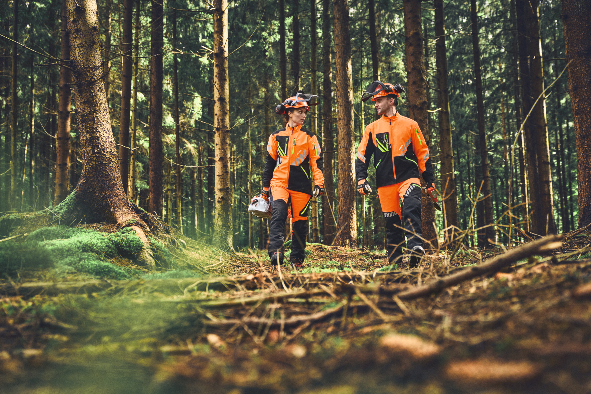 Two individuals walking in the forest wearing STIHL ADVANCE FlexTEC jacket and safety pants.