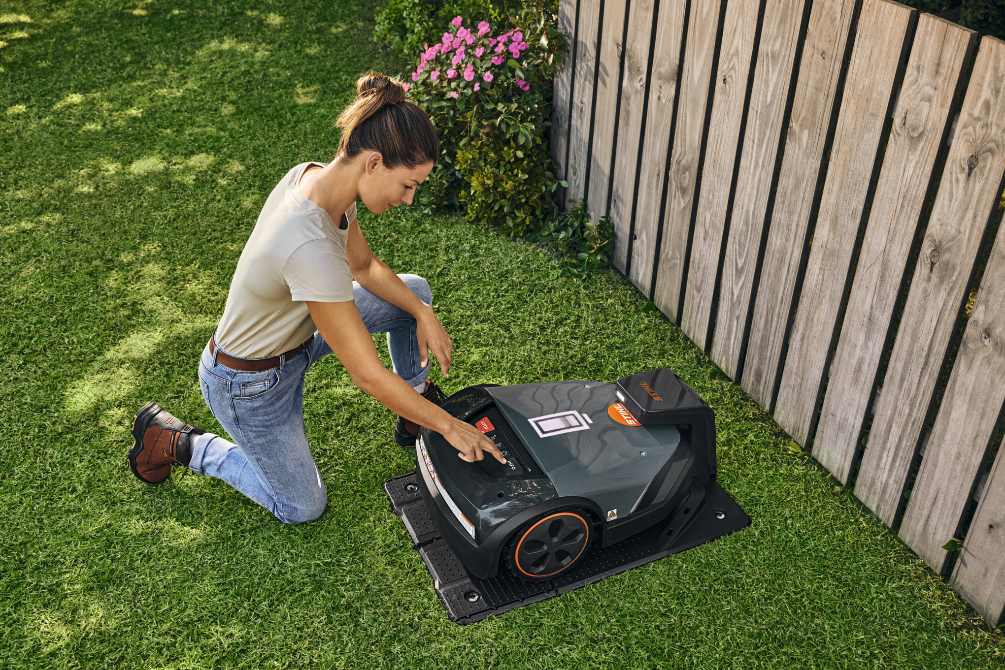 STIHL iMOW® robotic lawnmower in its docking station in the garden while a woman adjusts something on the robotic lawnmower.