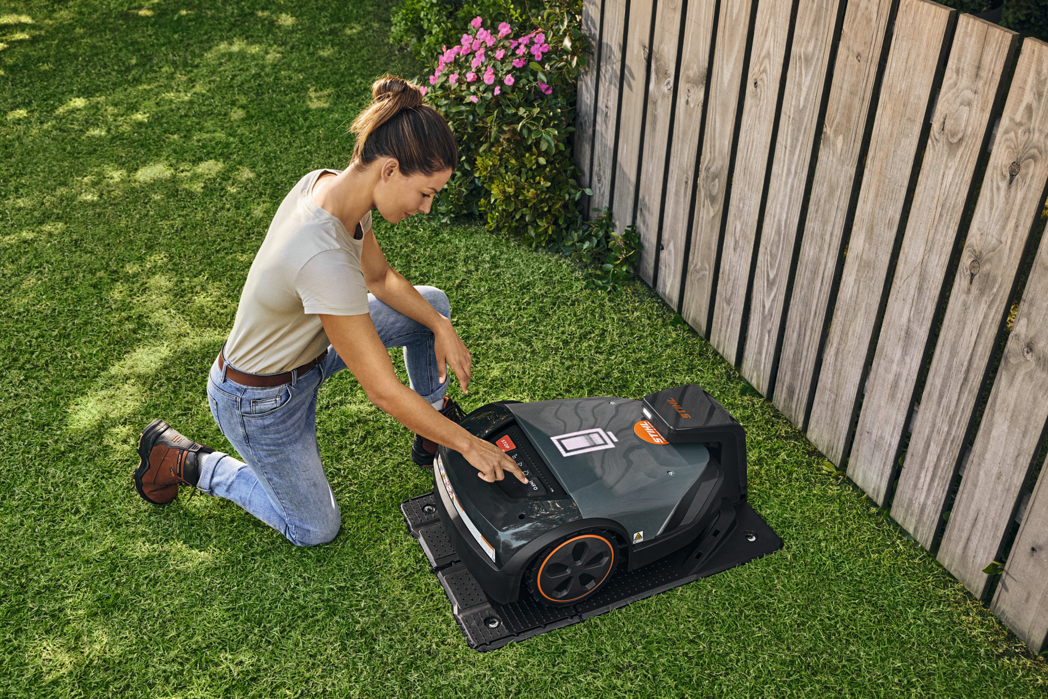 Robot de tonte STIHL iMOW® dans sa station d'accueil dans le jardin pendant qu'une femme règle quelque chose sur le robot de tonte.