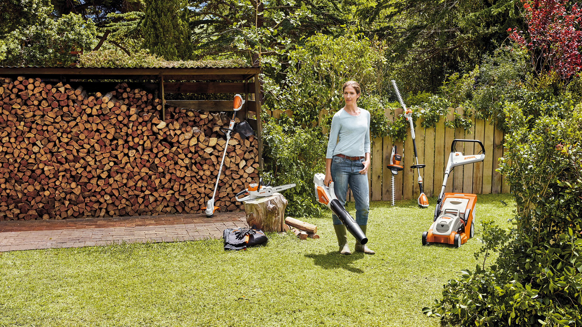 A woman standing in a garden, in front of a stack of wood with power tools from the AK-System 