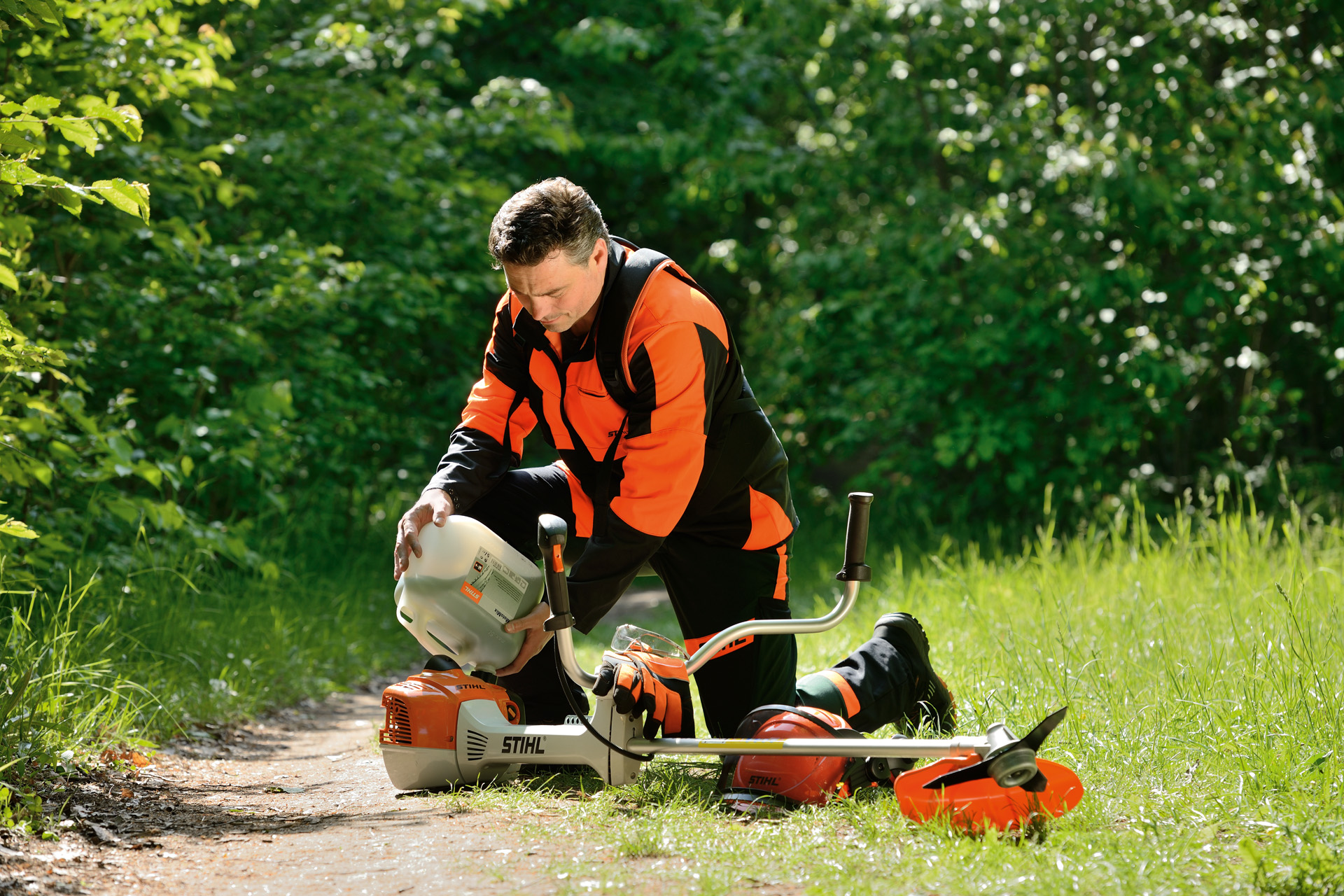 A man filling the fuel tank of a STIHL grass trimmer from a large bottle with shrubs behind him