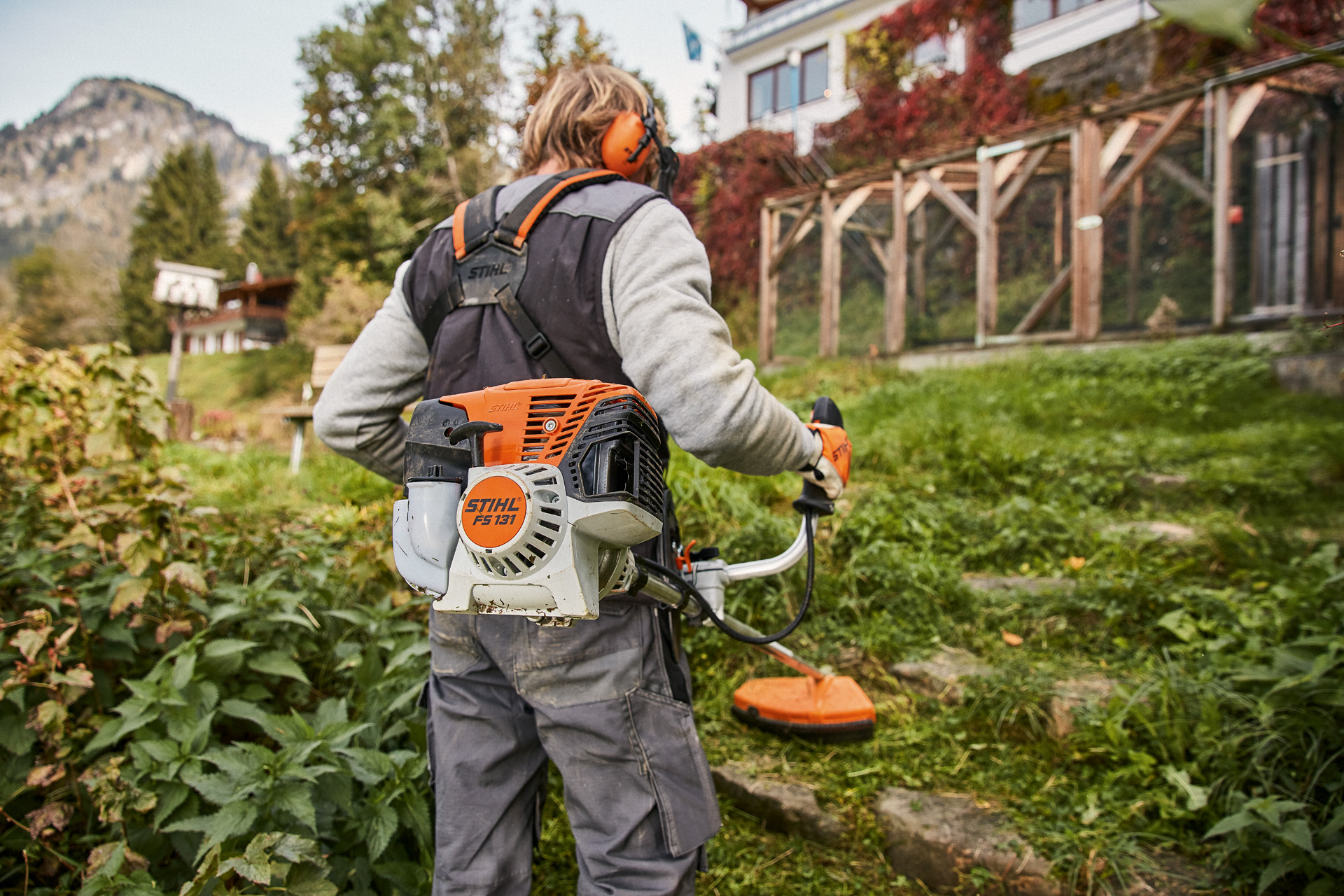 A person wearing protective gear using a STIHL FS 131 petrol brush cutter on uneven grassy ground in front of a house