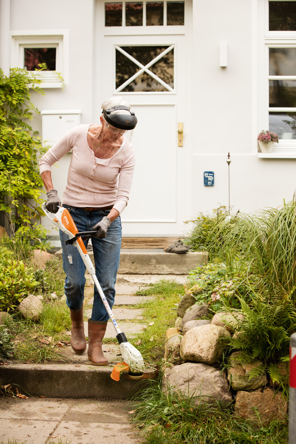 A woman wearing face protection using the STIHL FSA 45 cordless grass trimmer to  trim grass on a step in front of a house