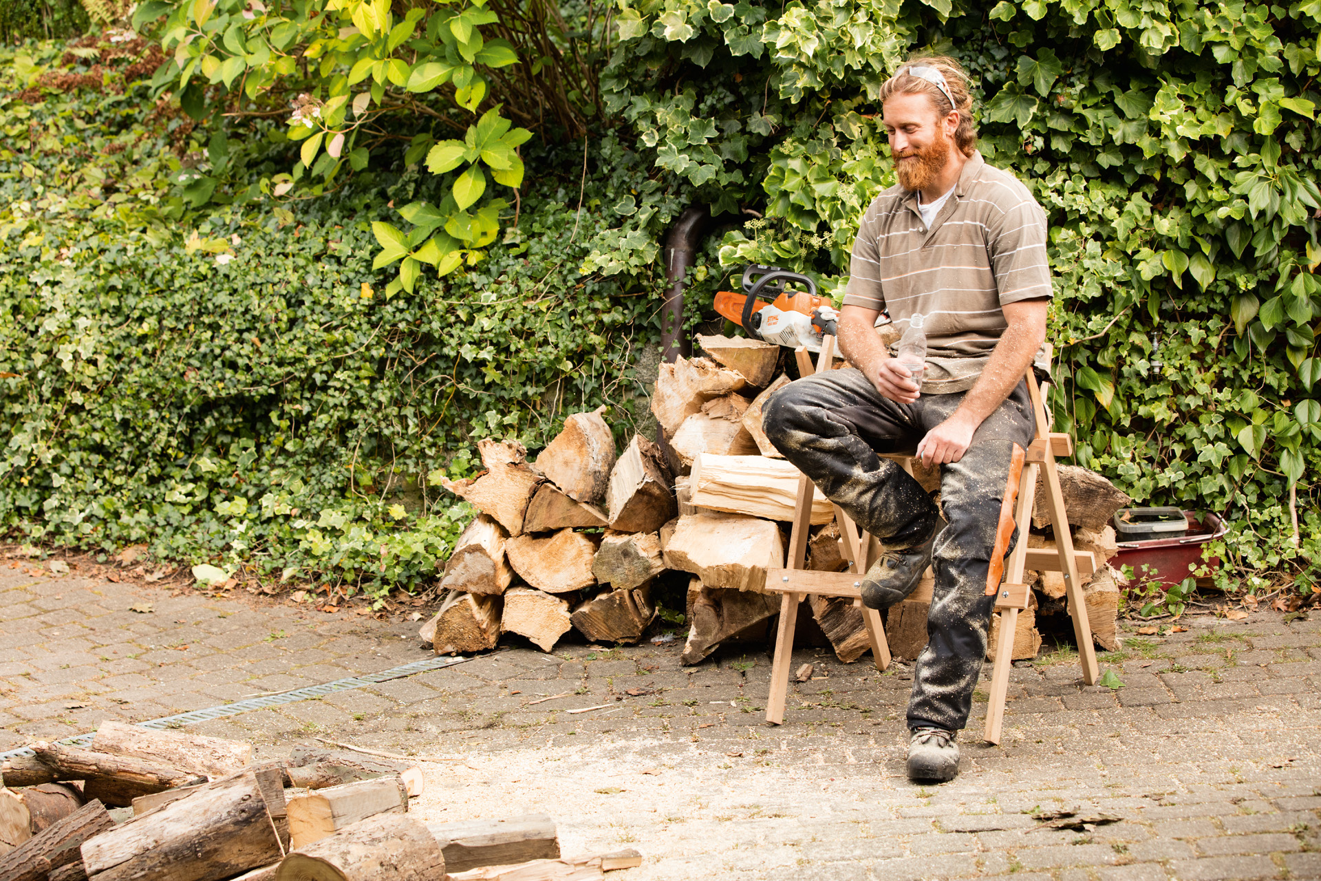Man sitting on a sawhorse in front of a pile of split logs.