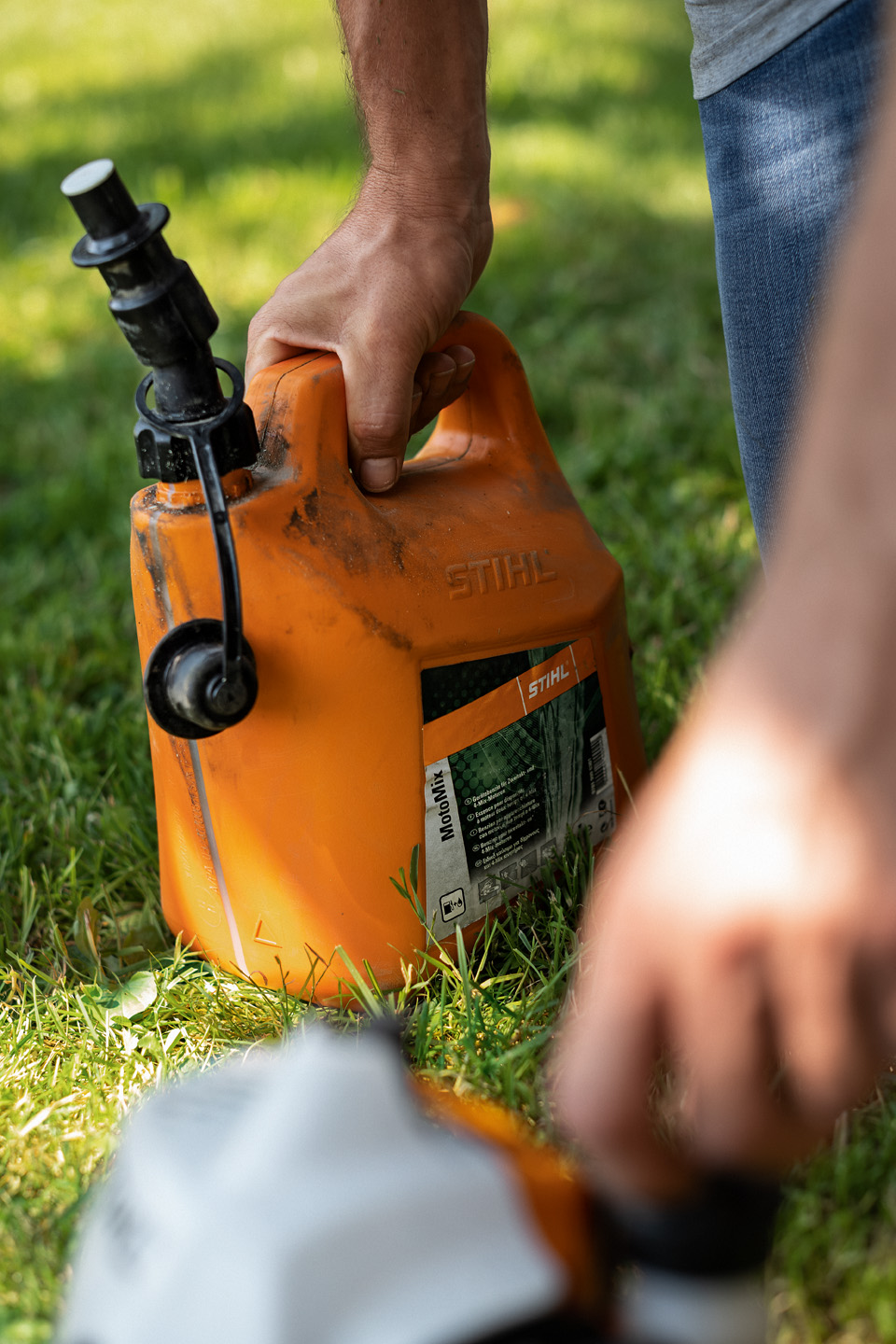 Un bidon sale de mélange de carburant STIHL MotoMix sur de l’herbe, avec une main ouvrant le bouchon du réservoir d’un outil au premier plan