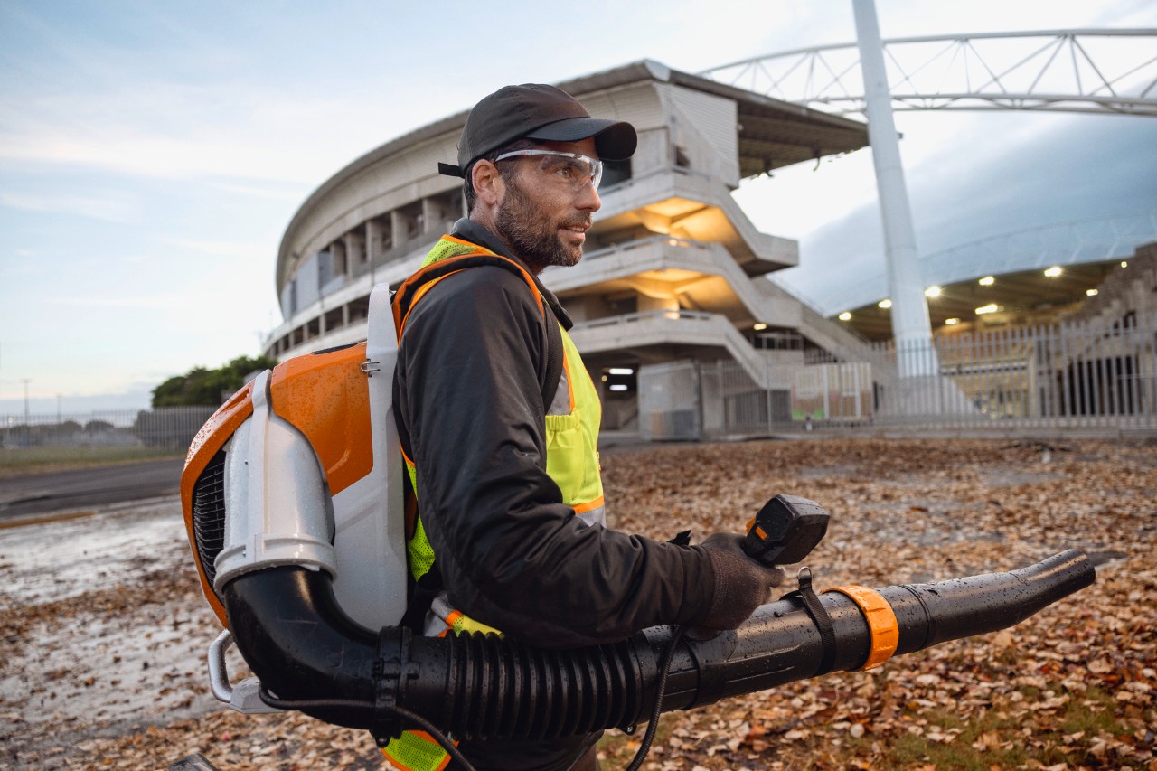 Late in the evening, a person is clearing leaves and debris from a stadium using the ALLPRO STIHL BRA 600 cordless leaf blower.