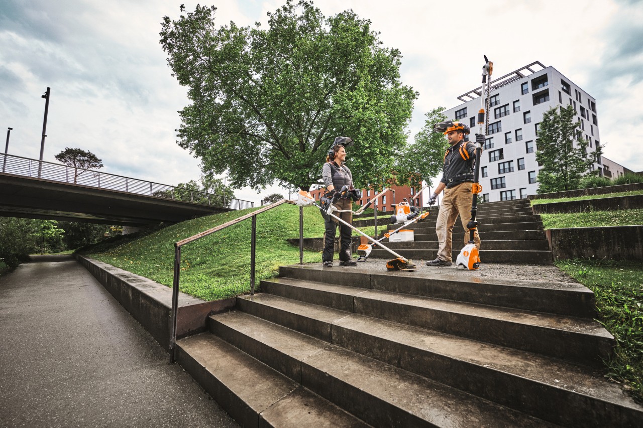 Two people with different devices from the ALLPRO battery system are standing on a staircase in front of some buildings.